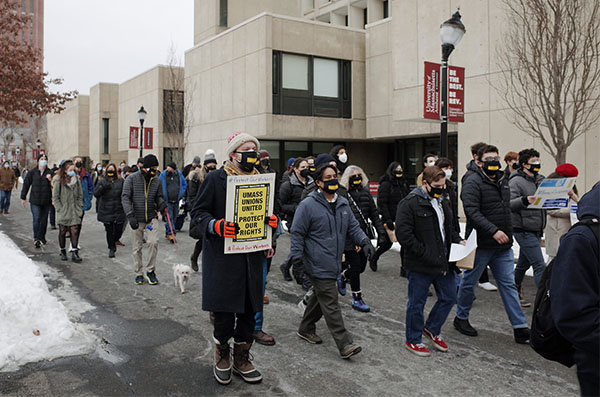 Union ralliers march past holding signs. One in front says umass unions united protect our rights